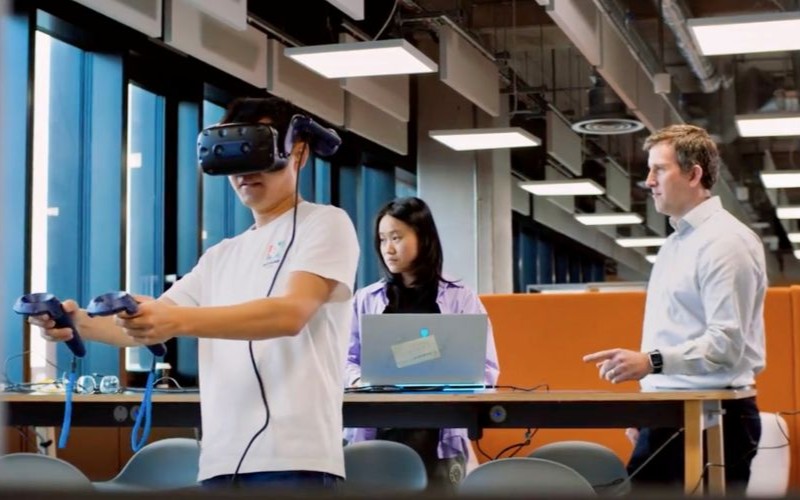 Student wearing a virtual reality headset with controllers, observed by others in a modern lab space