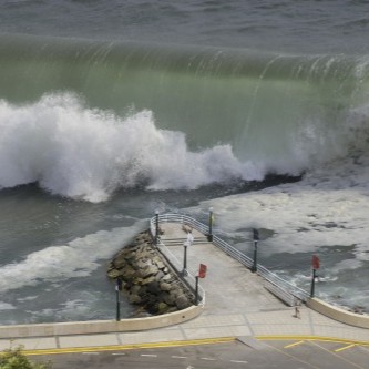 Large ocean wave crashing against a coastal seawall near a pedestrian walkway.