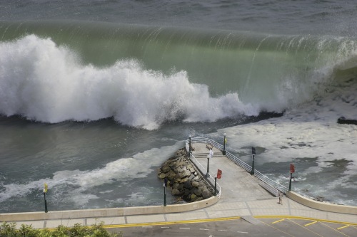Large ocean wave crashing against a coastal seawall near a pedestrian walkway.